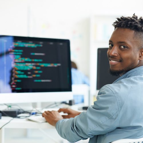 Happy guy looking at you while sitting by workplace and working with software